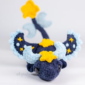 May include: A handmade crochet dragon in navy blue, light blue, and yellow. The dragon features star and moon accents on its wings and head, with a star-shaped horn and a star on the end of its tail. The dragon has large black eyes.