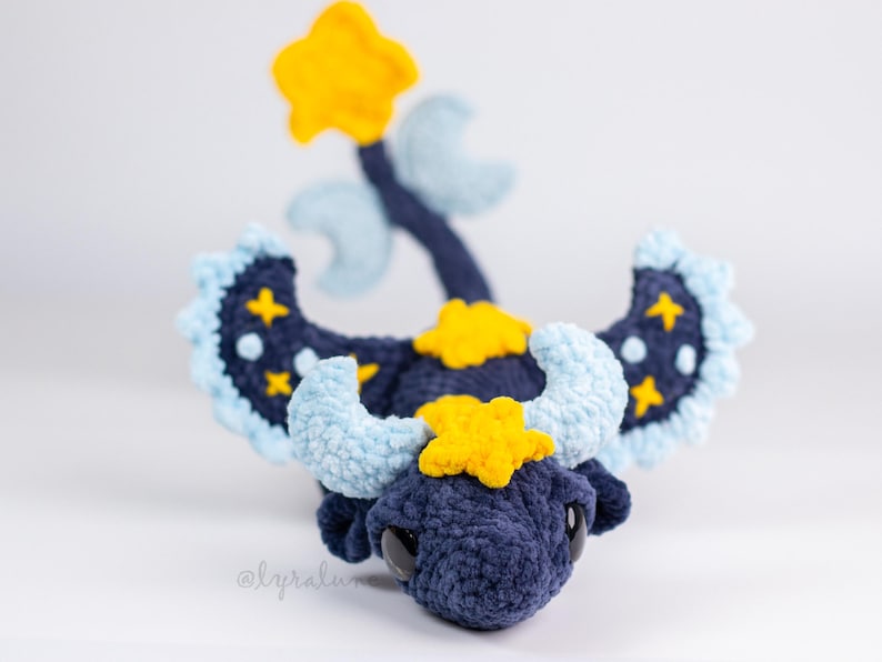 May include: A handmade crochet dragon in navy blue, light blue, and yellow. The dragon features star and moon accents on its wings and head, with a star-shaped horn and a star on the end of its tail. The dragon has large black eyes.
