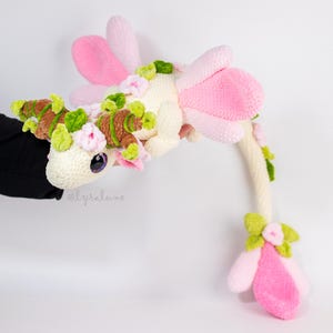 Puede incluir: Un peluche de dragón hecho a mano con ganchillo en crema, rosa, verde y marrón. El dragón presenta alas rosas, una cola larga y está adornado con detalles florales. El dragón se sostiene sobre un fondo blanco.
