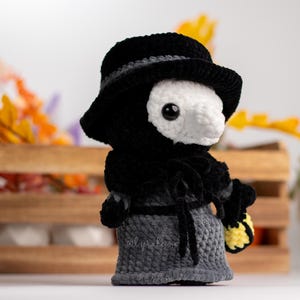 May include: A crocheted plush toy in the shape of a person wearing a black hat and a long black coat. The toy has a white face with a long beak and black eyes. The toy is holding a yellow and white crocheted object in its right hand.