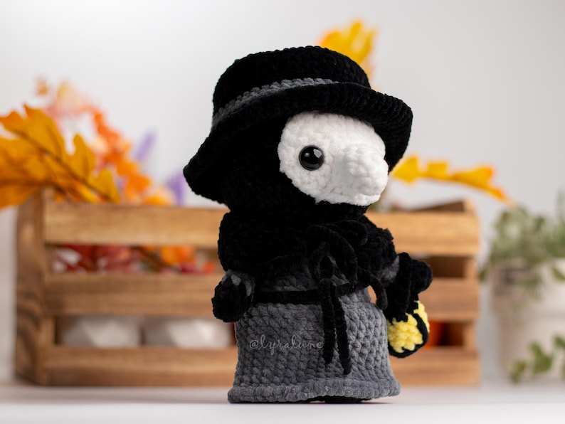 May include: A crocheted plush toy in the shape of a person wearing a black hat and a long black coat. The toy has a white face with a long beak and black eyes. The toy is holding a yellow and white crocheted object in its right hand.