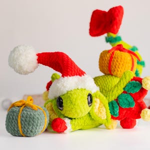 Puede incluir: Una criatura navideña de ganchillo con un gorro de Papá Noel rojo y blanco, cuerpo verde y detalles rojos. Acompañada de regalos de ganchillo en verde y amarillo, y un lazo rojo. Una decoración festiva.
