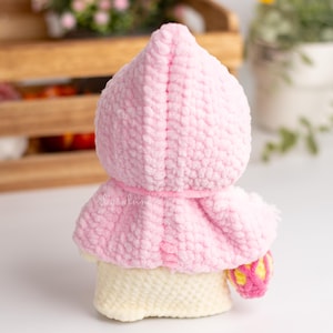 May include: A pink crocheted hooded doll with a white crocheted body and a yellow and pink crocheted accessory.