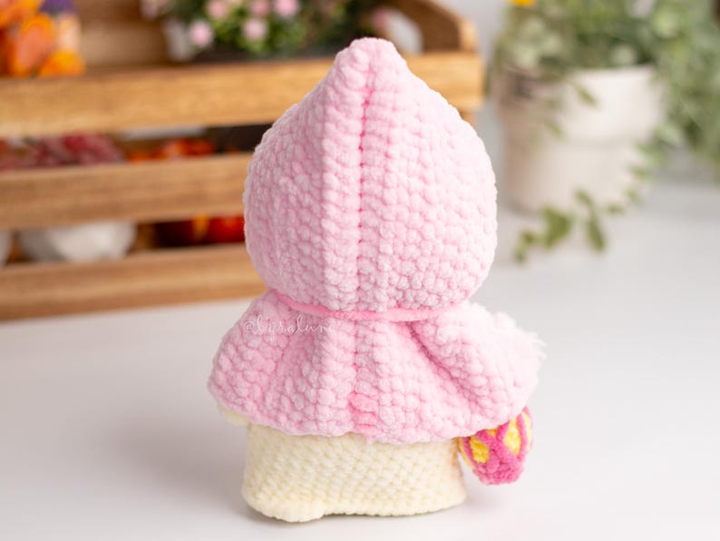 May include: A pink crocheted hooded doll with a white crocheted body and a yellow and pink crocheted accessory.