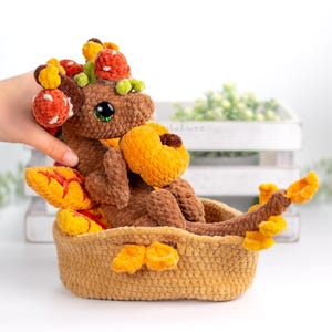 Peut inclure: Une créature crochetée marron avec de grands yeux verts, ornée d'accents orange et rouges, est assise dans un panier tressé. Le jouet en peluche a une forme de citrouille et est entouré de décorations sur le thème de l'automne. Le panier est également crocheté.