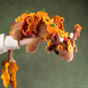 May include: A brown crocheted creature adorned with autumn decorations. It features orange pumpkins, leaves, and other seasonal elements. The plush toy has a long tail and is held by a person.