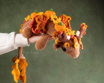 May include: A brown crocheted creature with fall-themed decorations. It features orange pumpkins, leaves, and other autumnal elements. The plush toy has a long tail and is held by a person.