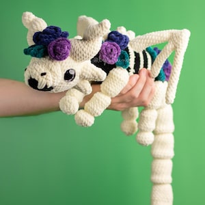 Op de afbeelding: Een witte gehaakte skeletdraak met een zwart-wit gestreept lichaam. De draak draagt ​​een kroon van paarse en turquoise bloemen.