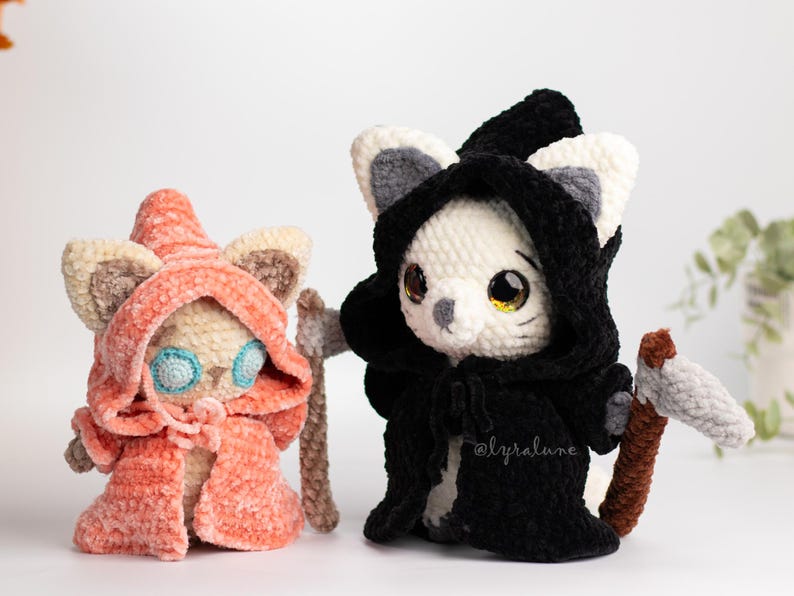 May include: Two crocheted cat plushies dressed in costumes. One is wearing a red hooded robe and the other is wearing a black hooded robe with a scythe.