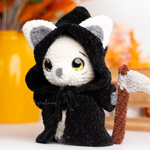 May include: A white crocheted cat wearing a black hooded robe and holding a grey crocheted scythe. The cat has large black eyes and a grey nose.