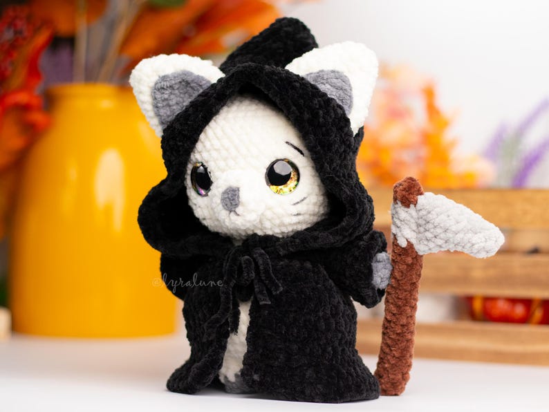 May include: A white crocheted cat wearing a black hooded robe and holding a grey crocheted scythe. The cat has large black eyes and a grey nose.
