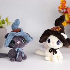 May include: Two plush cat figures, one gray and one white, each wearing a witch hat. The gray cat has a blue hat and bow, while the white cat has a brown hat and black bow. Both have large, iridescent eyes.