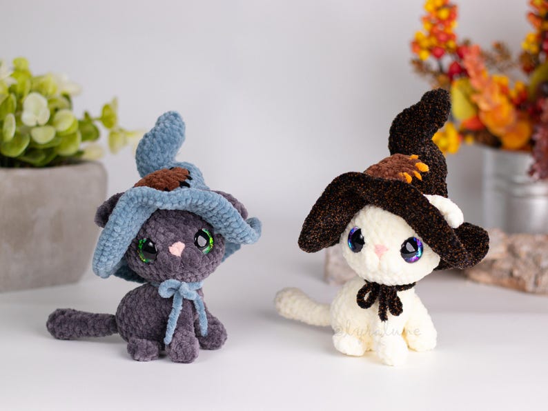 May include: Two plush cat figures, one gray and one white, each wearing a witch hat. The gray cat has a blue hat and bow, while the white cat has a brown hat and black bow. Both have large, iridescent eyes.