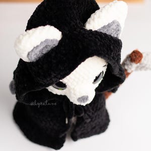 May include: A white crocheted animal wearing a black hooded cloak with a brown and white crocheted scythe. The animal has grey ears and black eyes.