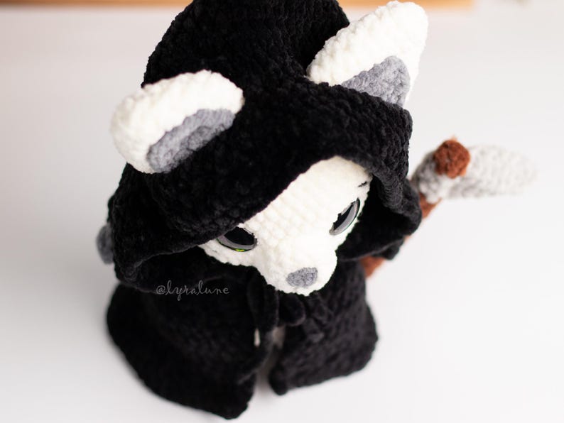 May include: A white crocheted animal wearing a black hooded cloak with a brown and white crocheted scythe. The animal has grey ears and black eyes.