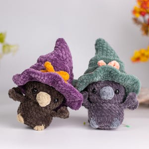 May include: Two handmade crochet plushies, each with a whimsical design. One is brown with a purple hat and orange bow, the other is purple with a green hat and pink bow. The plushies have small arms and legs.