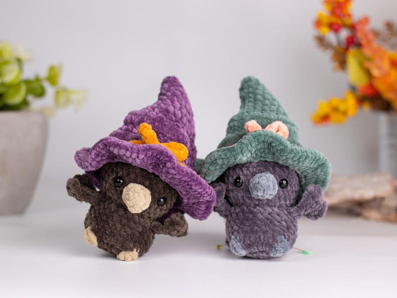 May include: Two handmade crochet plushies, each with a whimsical design. One is brown with a purple hat and orange bow, the other is purple with a green hat and pink bow. The plushies have small arms and legs.
