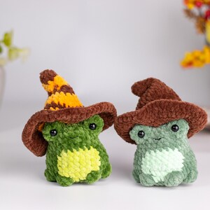 May include: Two crocheted frog figurines wearing brown witch hats. One frog is green with a yellow belly, the other is light green. The hats have a brown brim and a striped yellow and brown pointed top. The frogs have black eyes.