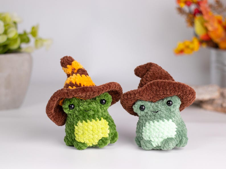 May include: Two crocheted frog figurines wearing brown witch hats. One frog is green with a yellow belly, the other is light green. The hats have a brown brim and a striped yellow and brown pointed top. The frogs have black eyes.