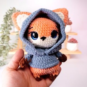 May include: A crocheted fox plush toy wearing a blue hooded sweatshirt. The fox has orange fur, white accents, and blue eyes.