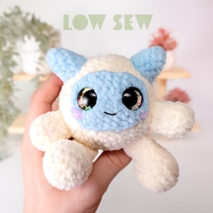 May include: A white and blue crocheted plush toy with large black eyes and a smiling mouth. The toy is being held in a person's hand. The text "LOW SEW" is visible in the background.