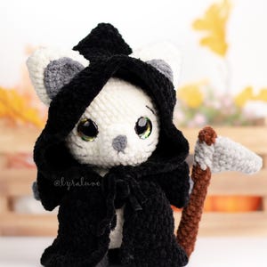 Könnte beinhalten: Eine weiße gehäkelte Katze, die einen schwarzen Kapuzenumhang trägt und eine graue gehäkelte Sense hält. Die Katze hat große grüne Augen und eine graue Nase. Die Katze sitzt auf einer Holzoberfläche mit einem braunen und orangen Hintergrund.