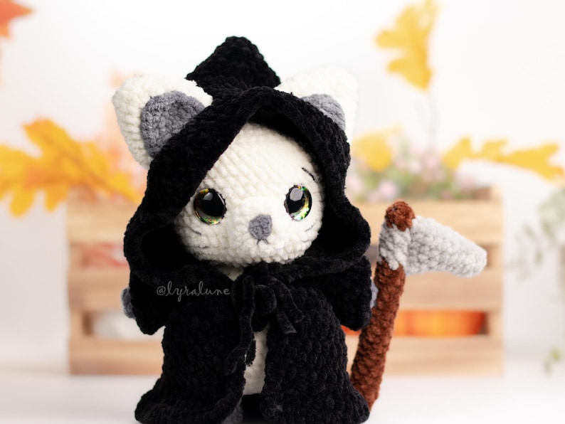May include: A white crocheted cat wearing a black hooded cloak and holding a grey crocheted scythe. The cat has large green eyes and a grey nose. The cat is sitting on a wooden surface with a brown and orange background.