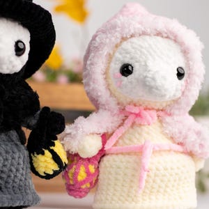 May include: Two crocheted plush toys, one in black with a witch's hat and the other in pink and white with a hooded cloak. Both toys are holding crocheted baskets.