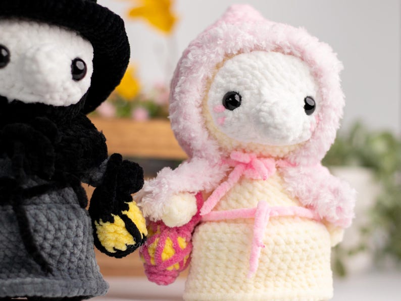 May include: Two crocheted plush toys, one in black with a witch's hat and the other in pink and white with a hooded cloak. Both toys are holding crocheted baskets.