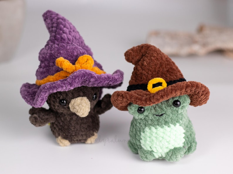 May include: Two handmade crochet plushies: a brown bird and a green frog, both wearing witch hats. The bird's hat is purple with an orange bow, and the frog's hat is brown with a yellow buckle. These whimsical toys are perfect for Halloween.