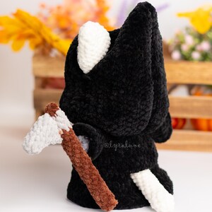 May include: Black crocheted plush toy with a white hood and tail, holding a brown and white crocheted axe. The toy is standing on a white surface.