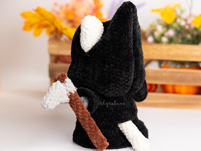 May include: Black crocheted plush toy with a white hood and tail, holding a brown and white crocheted axe. The toy is standing on a white surface.