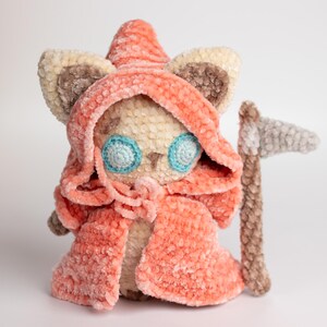 May include: A crocheted plush toy cat wearing a pink hooded cloak and holding a grey crocheted scythe. The cat has blue eyes and a beige body.