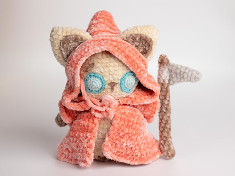 May include: A crocheted plush toy cat wearing a pink hooded cloak and holding a grey crocheted scythe. The cat has blue eyes and a beige body.