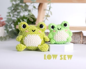 May include: Two crocheted green frog plushies with white accents. The larger frog is sitting on a white surface with the text "LOW SEW" in yellow.
