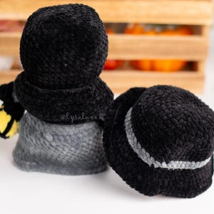 May include: Two black crocheted bucket hats with a gray trim. One hat is on top of a gray crocheted snowman with a yellow scarf.