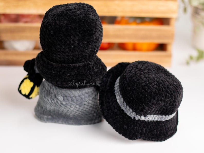 May include: Two black crocheted bucket hats with a gray trim. One hat is on top of a gray crocheted snowman with a yellow scarf.