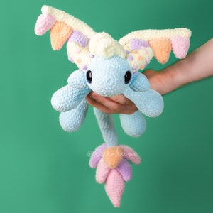 May include: A handmade crochet dragon plushie in shades of blue, pink, orange, and yellow. The dragon has large, round eyes and a long tail with a flower-like design. The wings are multi-colored. The plushie is held in a person's hand against a green background.