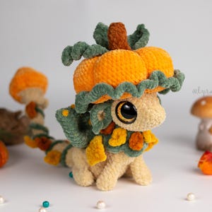 Puede incluir: Un peluche de dragón tejido a ganchillo con cuerpo color crema, grandes ojos negros y un sombrero en forma de calabaza naranja y verde. El dragón presenta detalles de hojas verdes y naranjas en el cuello y la cola, con el texto @lyralune visible.