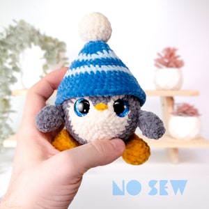 May include: A crocheted penguin wearing a blue and white striped hat with a white pom pom. The penguin has large black eyes and a yellow beak. The penguin is being held by a hand. The text "NO SEW" is visible in the bottom of the image.