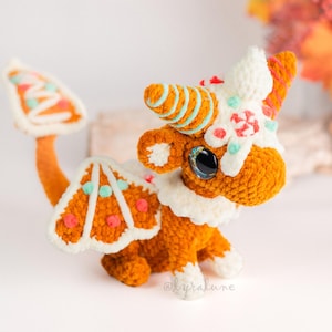 May include: A crocheted orange dragon with white and teal accents, resembling a gingerbread cookie. The dragon has large blue eyes, a white collar, and a tail with a gingerbread cookie design. The horns are decorated with teal stripes.