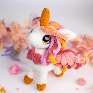 Puede incluir: Un unicornio blanco hecho a mano con ganchillo, con un cuerno dorado, ojos grandes y melena y alas coloridas. El unicornio tiene detalles en rosa, naranja y amarillo. El unicornio es un peluche.