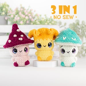 May include: Three crocheted mushroom plushies, each with a different coloured hat: burgundy, yellow, and teal. The plushies have white bodies and cute faces with black eyes and pink cheeks. The text "3 IN 1 - NO SEW -" is displayed above the plushies.