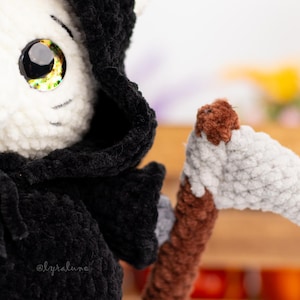 May include: A white crocheted cat wearing a black hooded robe and holding a brown and white crocheted scythe. The cat has large, yellow eyes.