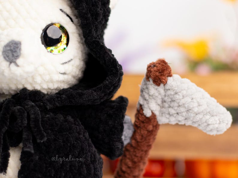 May include: A white crocheted cat wearing a black hooded robe and holding a brown and white crocheted scythe. The cat has large, yellow eyes.