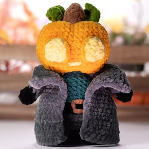 Puede incluir: Un juguete de peluche de calabaza tejido a crochet que lleva un abrigo gris y turquesa. La calabaza tiene una cara amigable con ojos amarillos y un tallo marrón.