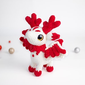 Op de afbeelding: Een handgemaakt, wit en rood gehaakt wezen met grote, gouden ogen, rode geweien en vleugels. De knuffel heeft rode accenten op de voeten en het lichaam. Decoratieve ornamenten in rood, goud en zilver zijn verspreid.