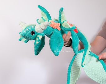 Peut inclure: Une peluche de dragon crochetée en vert d'eau et vert menthe, avec de grandes ailes et une longue queue. Le dragon a des cornes blanches, une petite étoile de mer orange et un accent rouge. Le dragon est tenu par une personne.