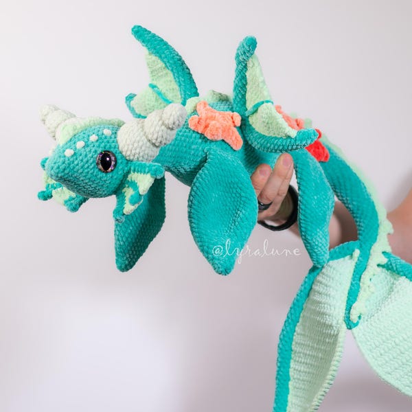 Op de afbeelding: Een gehaakte drakenknuffel in zeegroen en mintgroen met grote vleugels en een lange staart. De draak heeft witte hoorns, een kleine oranje zeester en een rood accent. De draak wordt door een persoon vastgehouden.