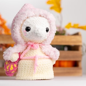 May include: A crocheted plush toy wearing a pink hooded cloak and holding a pink and yellow crocheted basket. The toy has a white body with black eyes and a pink nose.
